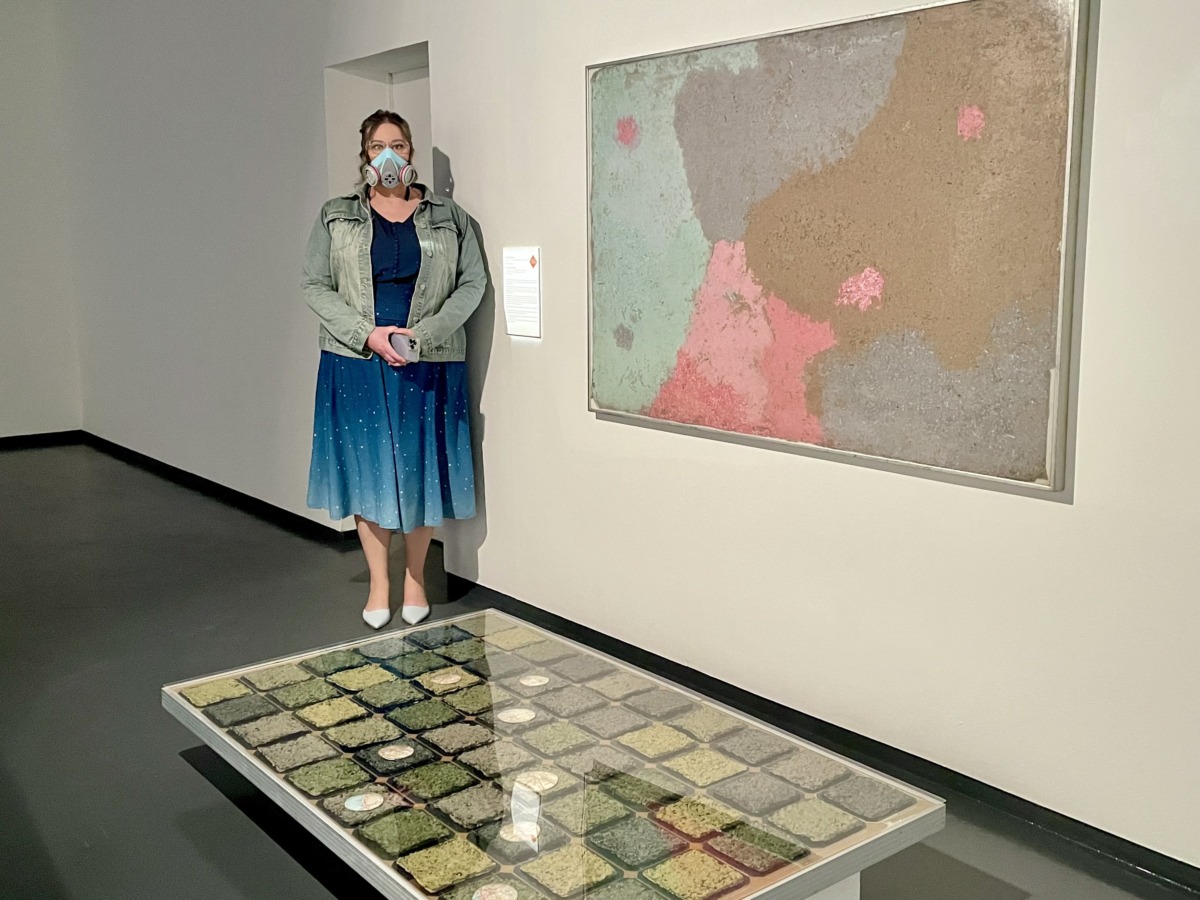 image of a woman in a blue dress and respirator in front of artworks made from solar panels